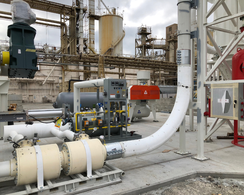 Flash Rockwell low energy industrial evaporator installed on a skid at an outdoor plant, with white piping and cylindrical components feeding a compact gray control skid in front of large process tanks and steel pipe racks.